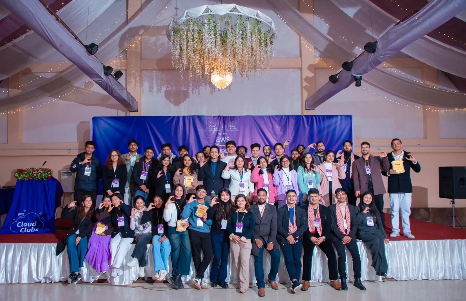 Large group of attendees posing on and in front of the stage in a decorated hall under a chandelier, many making hand gestures