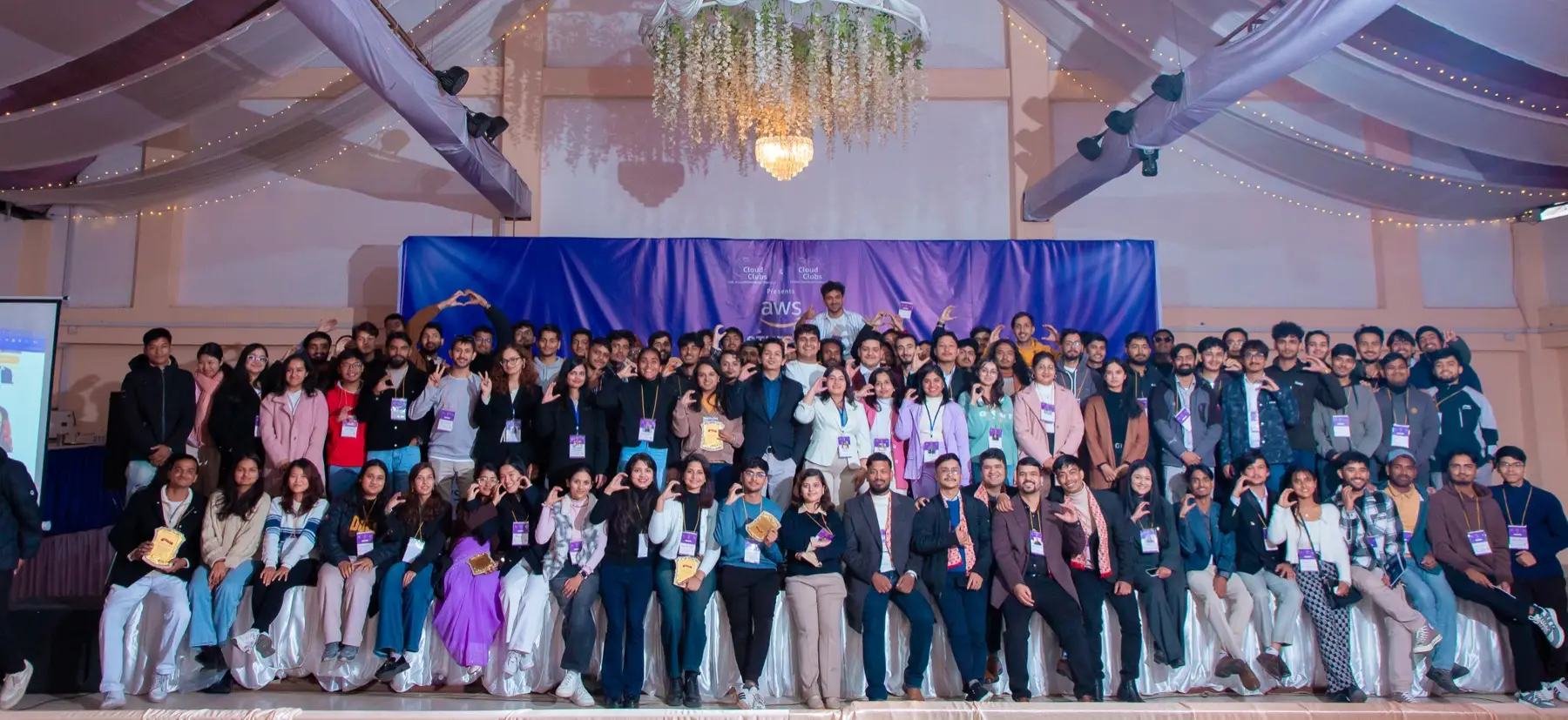 Wide shot of a packed crowd of attendees seated and standing in tiers in front of the event backdrop, with a chandelier above