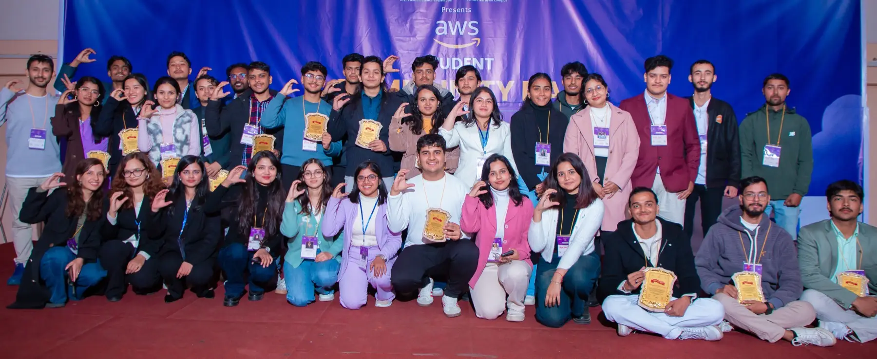 Large group of volunteers and participants, some kneeling and some standing, posing with plaques on a red carpet in front of the event banner