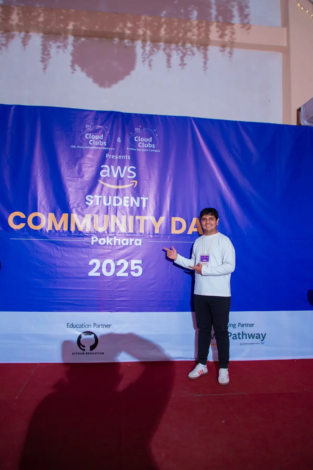 Person in a white sweater gesturing toward the AWS Student Community Day Pokhara 2025 backdrop on a red-carpeted stage
