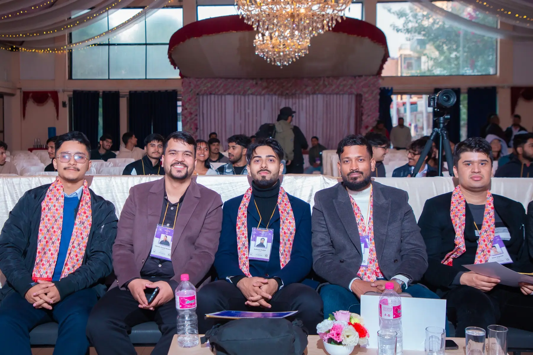 Five men with traditional scarves and badges sitting in a row in the audience, with the hall and a chandelier behind them