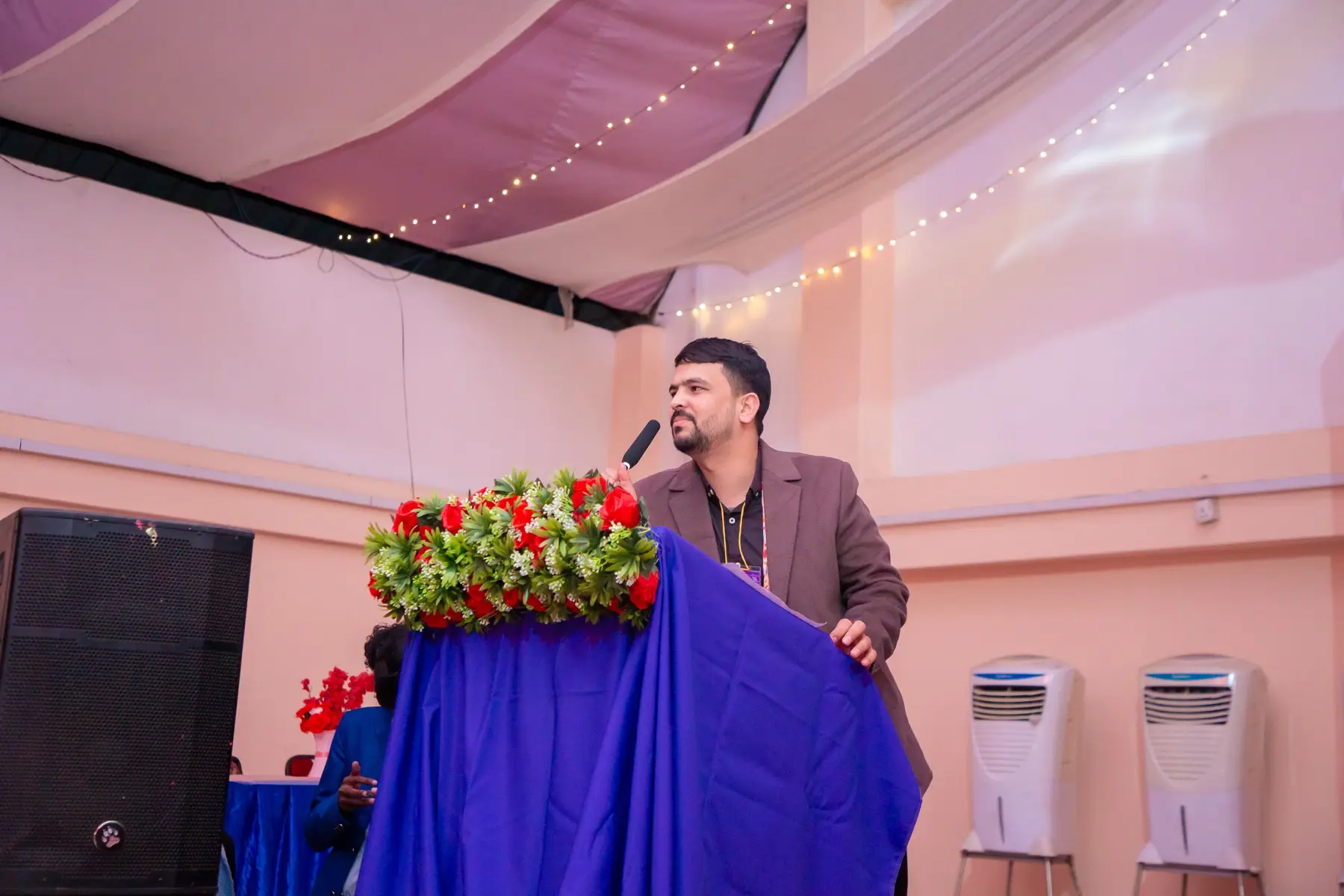 Speaker in a brown blazer talking at a podium with a flower arrangement and blue cloth