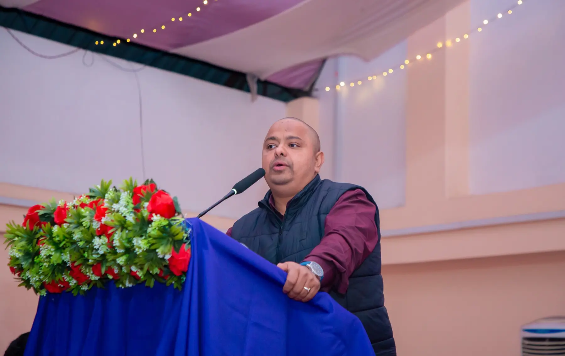 Speaker in a dark vest speaking into a microphone at a flower-decorated podium