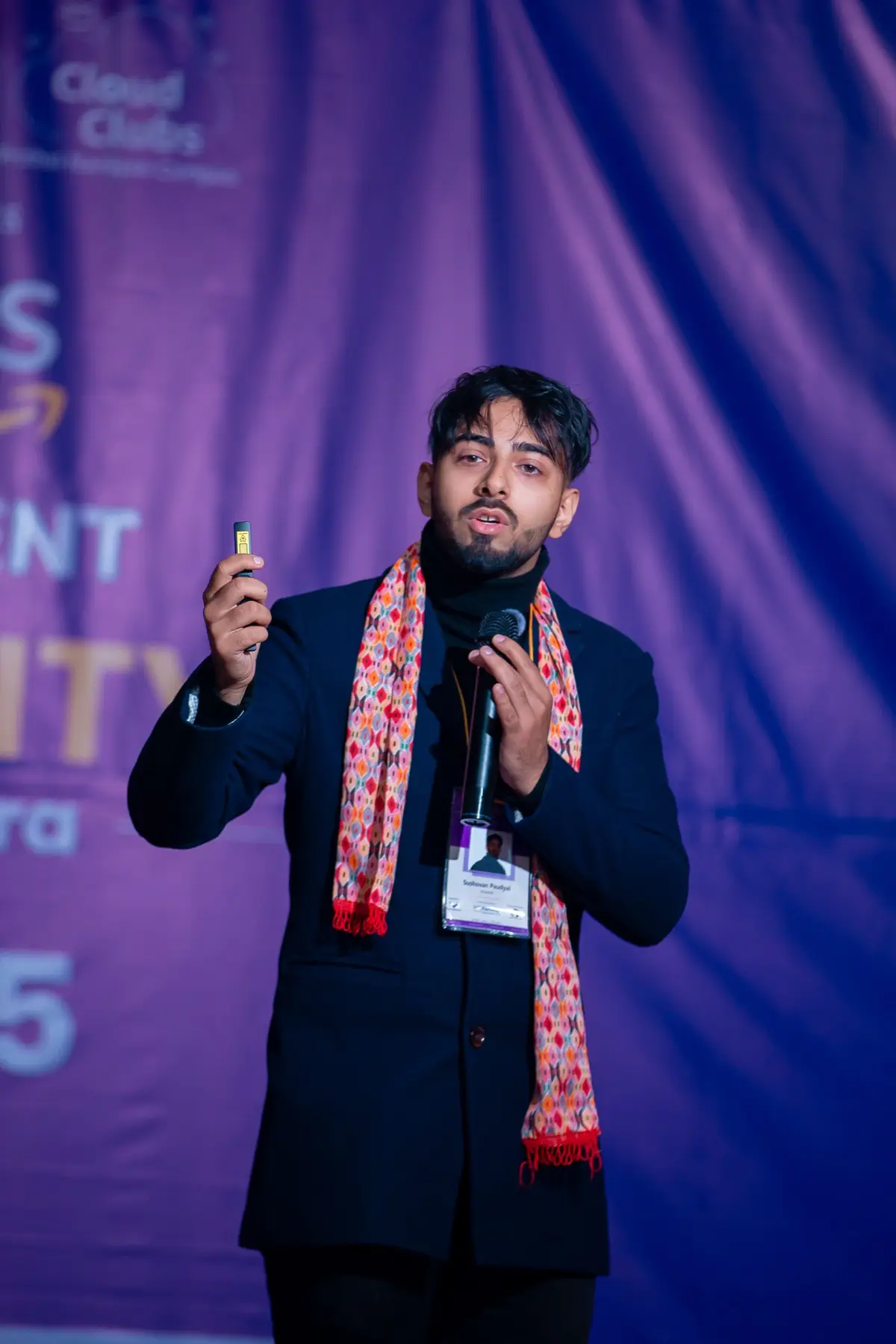 Speaker with a patterned scarf holding a microphone and a clicker in front of the purple event backdrop