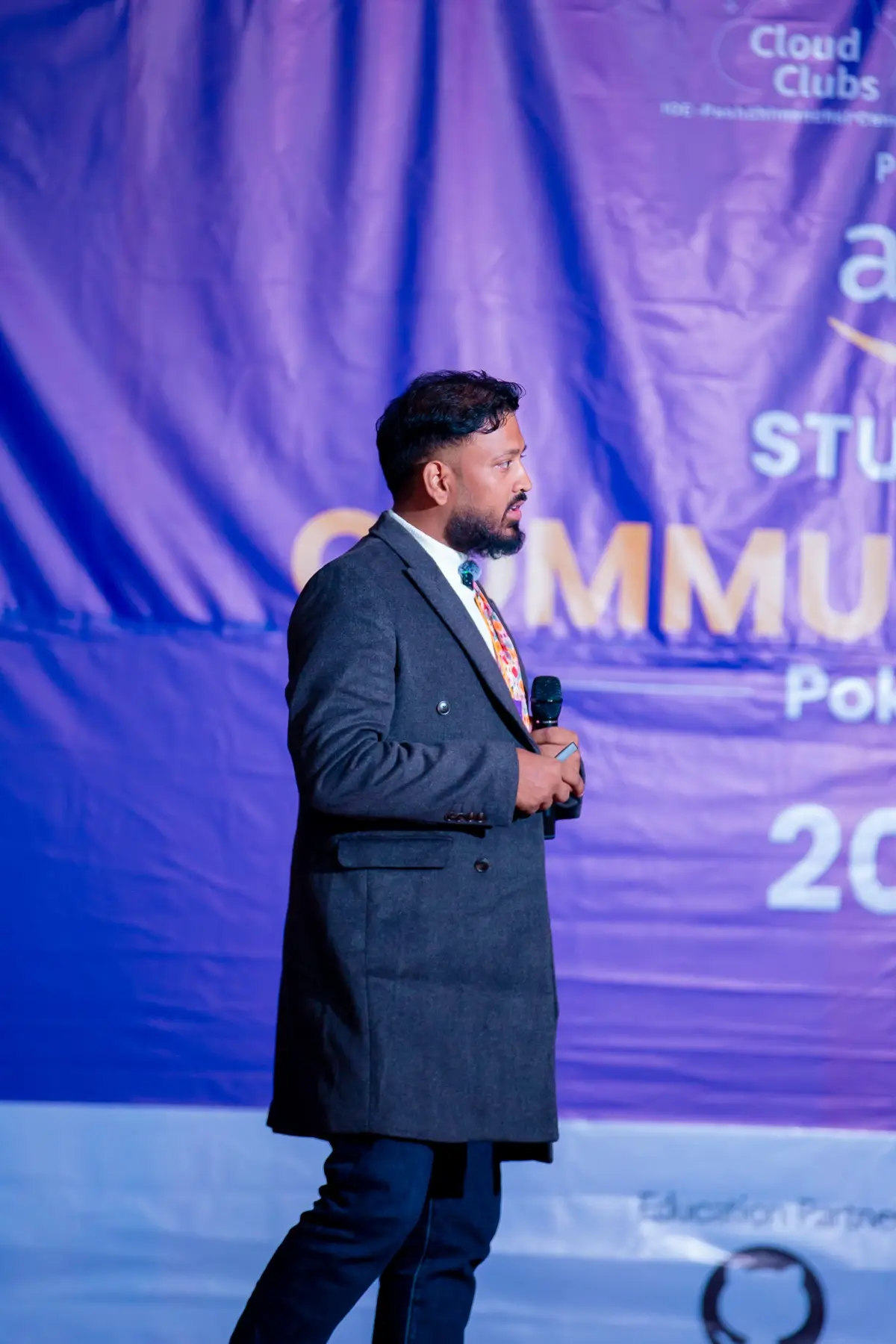 Speaker in a long grey coat holding a microphone and walking across the stage in front of the event backdrop